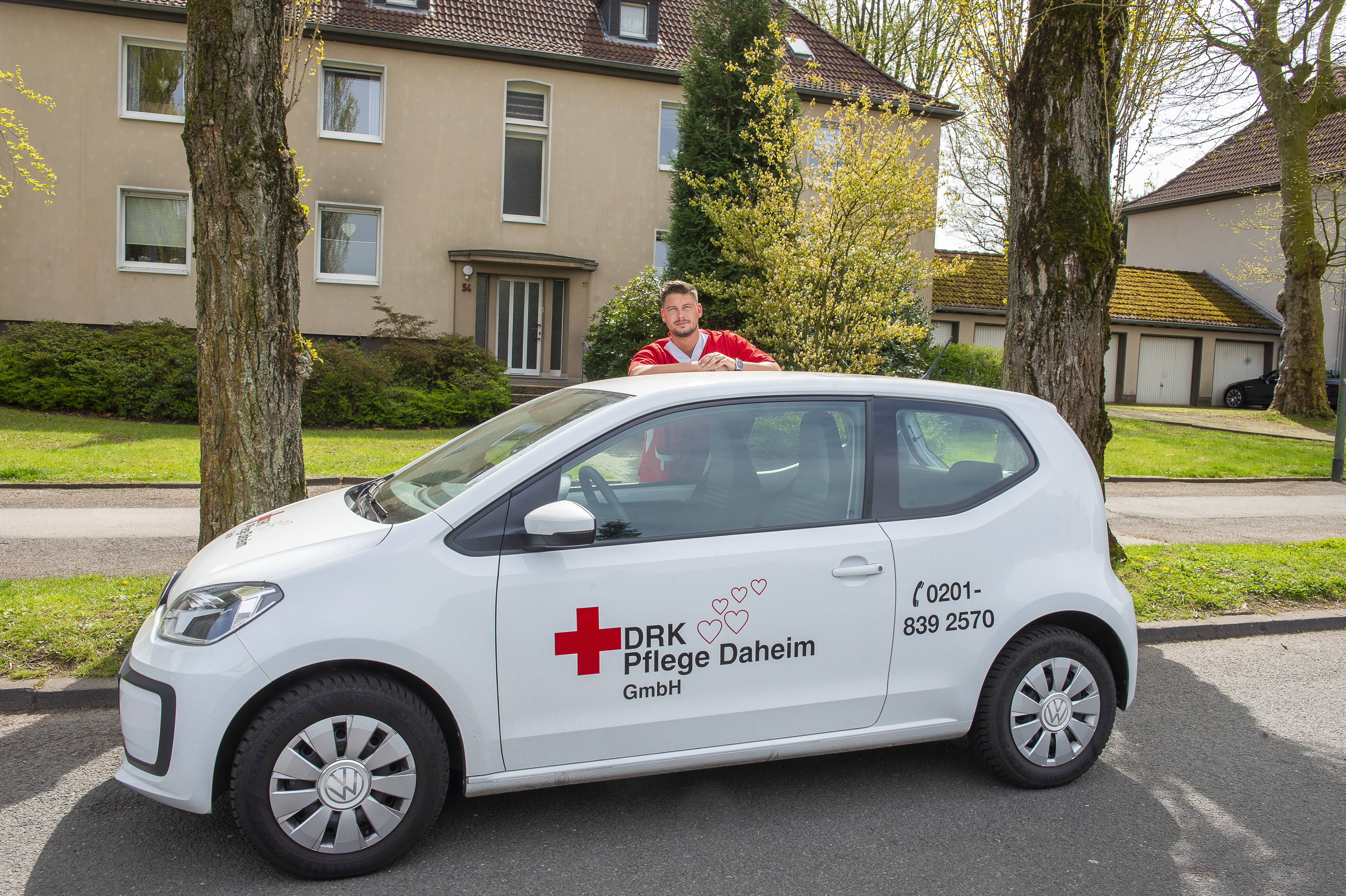 Ein Mitarbeiter in rotem Kasack steht hinter dem weißen Auto der DRK Essen Pflege Daheim und schaut über das Autodach.