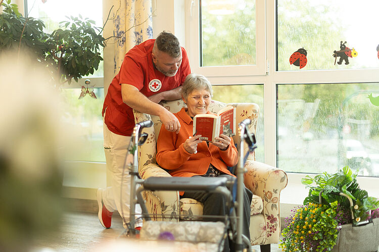 Pflege mit Herz Eine männliche Pflegekraft beugt sich von hinten über einen großen Ohrensessel indem eine ältere Bewohnerin ein Buch liest.