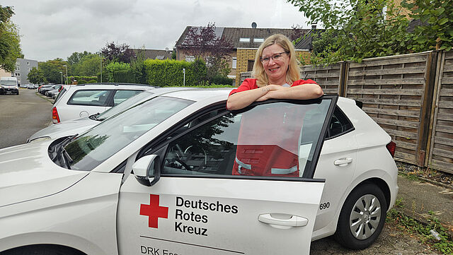 Mitarbeiterin des DRK-Pflegedienstes „Pflege Daheim“ steht an einem Dienstfahrzeug in einem Wohngebiet.