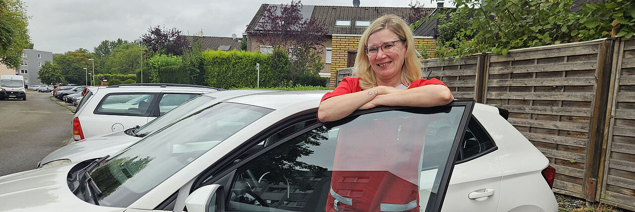Pflege Daheim – Ambulante Pflege des DRK Essen Mitarbeiterin des DRK-Pflegedienstes „Pflege Daheim“ steht an einem Dienstfahrzeug in einem Wohngebiet.