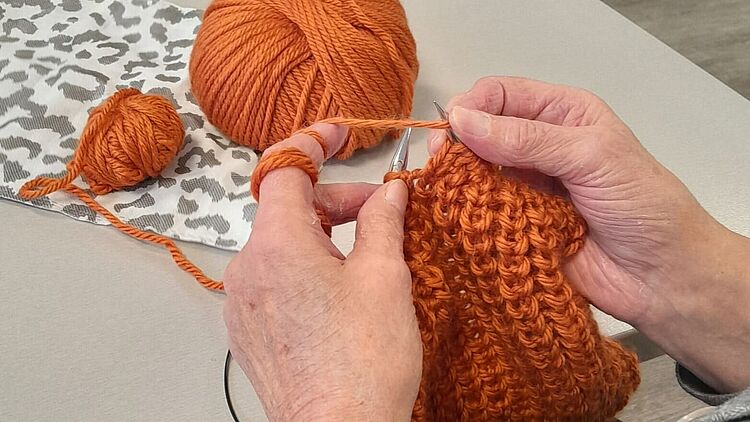 Strickwerkstatt Hände stricken mit orangefarbener Wolle. Im Hintergrund sind orangene Wollknäule zu sehen.