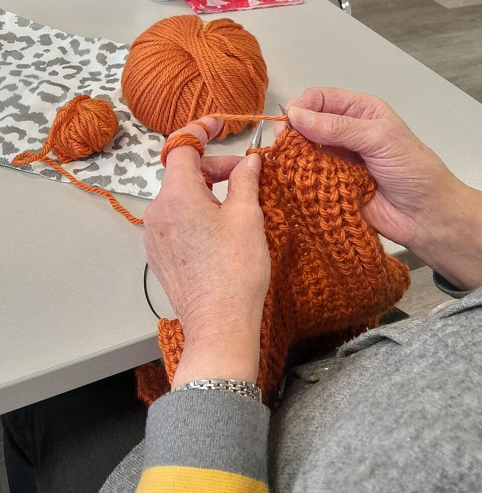 Strickwerkstatt Hände stricken mit orangefarbener Wolle. Im Hintergrund sind orangene Wollknäule zu sehen.