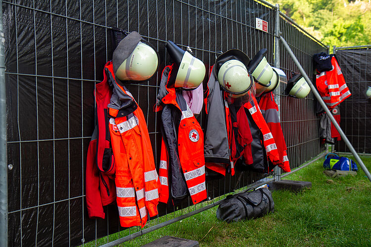 Einsatzkleidung und mehrere Helme hängen nebeneinander an einem Bauzaun.