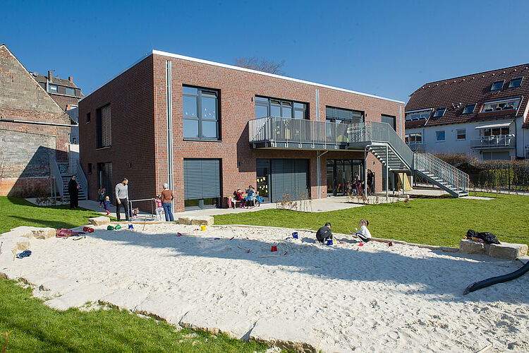 Das braune moderne Backsteingebäude der Kita Claras Garten mit Garten und Sandkasten im Vordergrund, in dem mehrere Kinder spielen.
