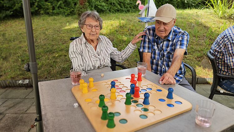 Eine Seniorin und ein Senior spielen Mensch-ärgere-dich-nicht an einem Tisch im Garten. Sie legt ihm eine Hand auf die Schulter.