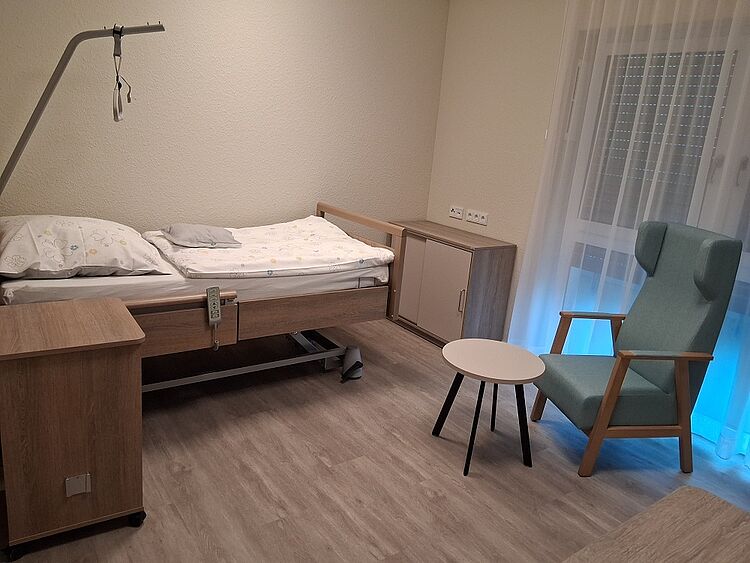 Einzelzimmer Ein Pflegebett steht vor einer hellen Wand, schräg dahinter steht ein Ohrensessel mit einem Beistelltisch vor einem Bodentiefen Fenster.
