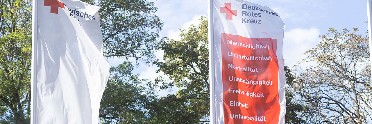 Drei Fahnen des DRK wehen im Wind. Auf einer sind die Grundsätze auf rotem Hintergrund geschrieben.