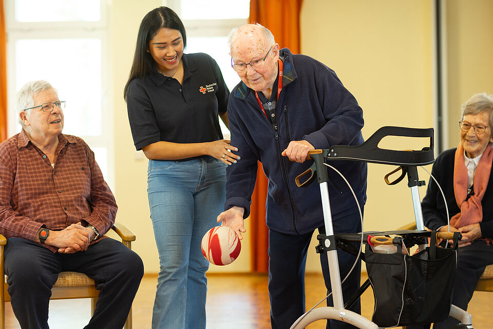Freizeit im Seniorenzentrum Rüttenscheid Eine Sozialarbeiterin unterstützt einen Bewohner mit Rollator dabei eine große Kugel zu werfen. Daneben sitzen weitere ältere Bewohner auf Stühlen und schauen zu.