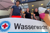 Ein Ehrenamtlicher der Wasserwacht Essen präsentiert mit der Inhaberin und einer Mitarbeiterin eines Eiscafes den Start der Sommeraktion.