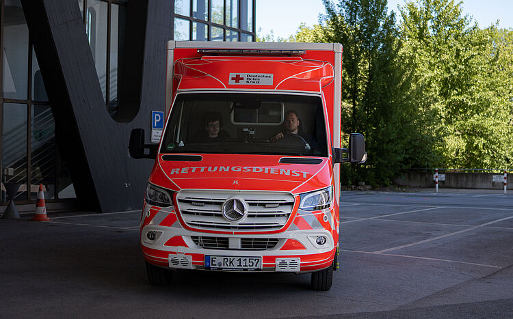 Rettungswagen steht draußen im Hintergrund ein graues Gebäude und Bäume.