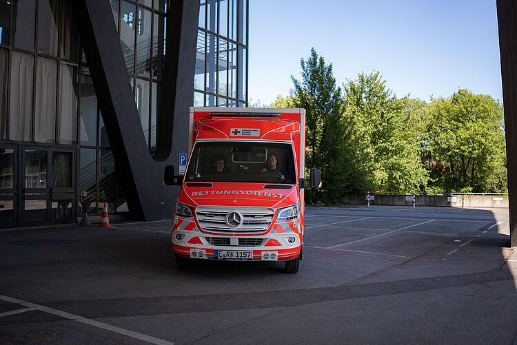 Rettungswagen steht draußen im Hintergrund ein graues Gebäude und Bäume.