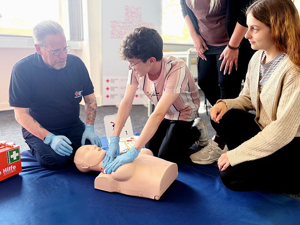 Erste Hilfe In einem Erste-Hilfe-Kurs demonstriert ein Ausbilder gemeinsam mit einer teilnehmenden Person die Herzdruckmassage an einer Reanimationspuppe. Während die Person die Wiederbelebungsmaßnahme durchführt, beobachtet der Ausbilder aufmerksam und gibt Unterstützung. Zwei weitere Teilnehmende verfolgen die Übung. Im Vordergrund steht ein Erste-Hilfe-Kasten, im Hintergrund sind Schulungsmaterialien zu sehen.