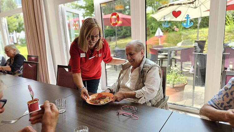 Eine Seniorin sitzt an einer gedeckten Tafel. Eine Mitarbeiterin des DRK Essen reicht ihr einen gefüllten Teller mit Mittagessen. Im Hintergrund ist durch große Glasscheiben ein Garten zu sehen.