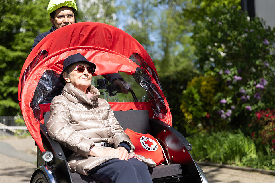Ausflug mit der Rikscha Eine ältere Frau mit Sonnenhut und Sonnenbrille sitzt lächelnd in einer roten Fahrradrikscha. Der Fahrer trägt einen neungelben Helm.
