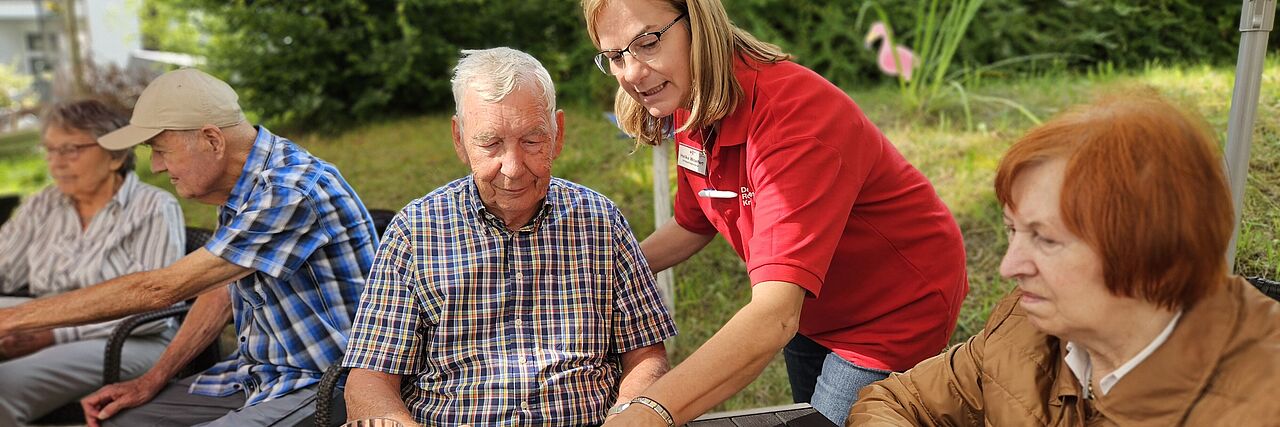 Ein älterer Herr sitzt im Garten an einem Tisch mit einem Brettspiel. Eine Pflegekraft zeigt auf eine Spielfigur. Rechts sitzt eine Seniorin, im Hintergrund unterhalten sich zwei Senioren.