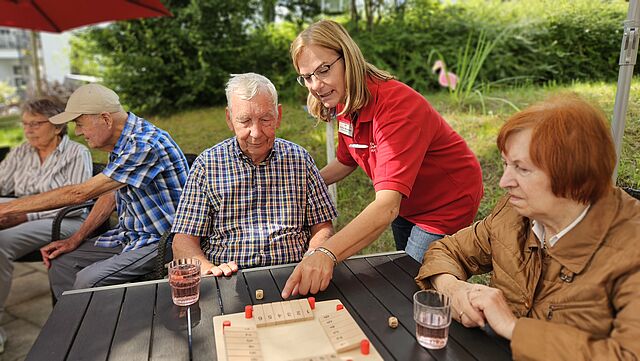 Ein älterer Herr sitzt im Garten an einem Tisch mit einem Brettspiel. Eine Pflegekraft zeigt auf eine Spielfigur. Rechts sitzt eine Seniorin, im Hintergrund unterhalten sich zwei Senioren.