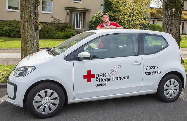 Ein Mitarbeiter in rotem Kasack steht hinter dem weißen Auto der DRK Essen Pflege Daheim und schaut über das Autodach.