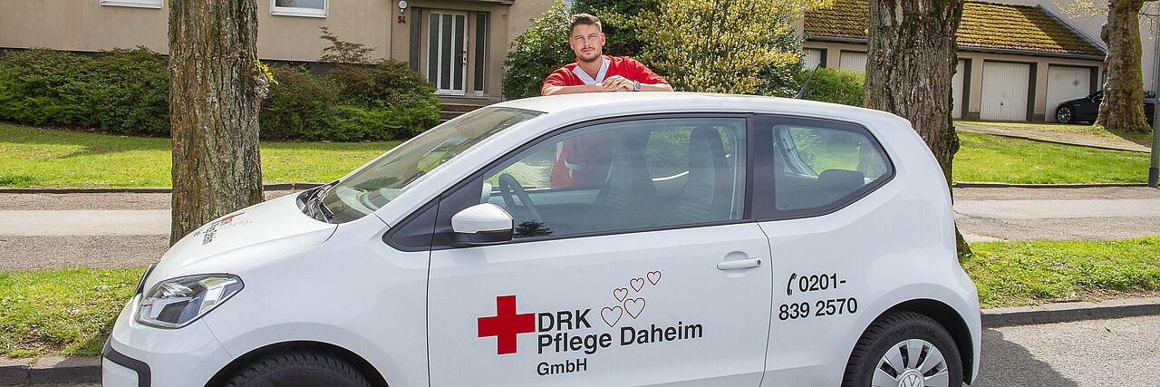 Ein Mitarbeiter in rotem Kasack steht hinter einem weißen Auto der DRK Essen Pflege Daheim. Er schaut über das Autodach.