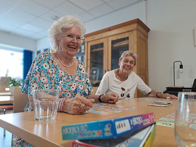 Zwei ältere Frauen spielen am Tisch ein Gesellschaftsspiel. Beide lächeln.
