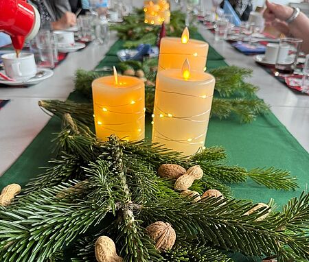 Es sind Tannenzweige und drei brennende Kerzen als Tischdekoration an einer gedeckten Tafel zu sehen.