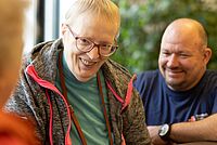 Eine Bewohnerin des Pflegezentrums schaut lachend in Richtung Kamera. Ein Mitarbeitender des Roten Kreuzes ist unscharf hinter ihr zu erkennen.