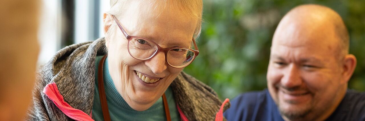Eine Bewohnerin des Pflegezentrums schaut lachend in Richtung Kamera. Ein Mitarbeitender des Roten Kreuzes ist unscharf hinter ihr zu erkennen.