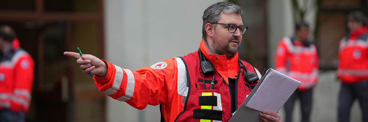Ein Mann mit DRK-Einsatzkleidung aus den Bereitschaften zeigt mit ausgestrecktem Arm in eine Richtung und liest währenddessen auf einem Zettel etwas.