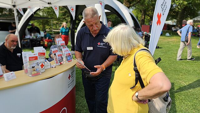Ein Mann in DRK-Shirt erklärt einer älteren Frau in gelben T-Shirt etwas vor einem DRK-Infostand.