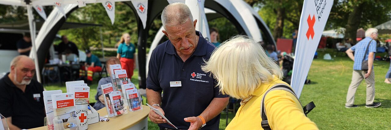 Ein Mann in DRK-Shirt erklärt einer älteren Frau in gelben T-Shirt etwas vor einem DRK-Infostand.