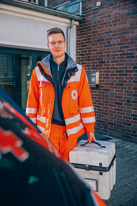 Junger Mann in DRK-Einsatzkleidung trägt einen grauen Transportkoffer in der Hand.