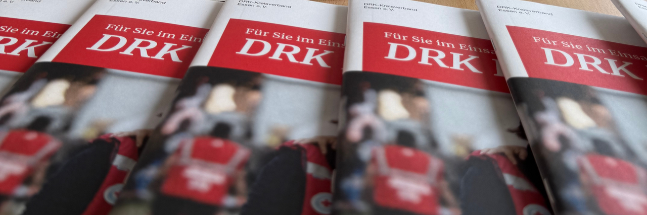 Aktuelle Ausgabe der DRK Essen-Broschüre „Für Sie im Einsatz.“ in einem Aufsteller auf einem Konferenztisch, umgeben von mehreren ausliegenden Exemplaren.