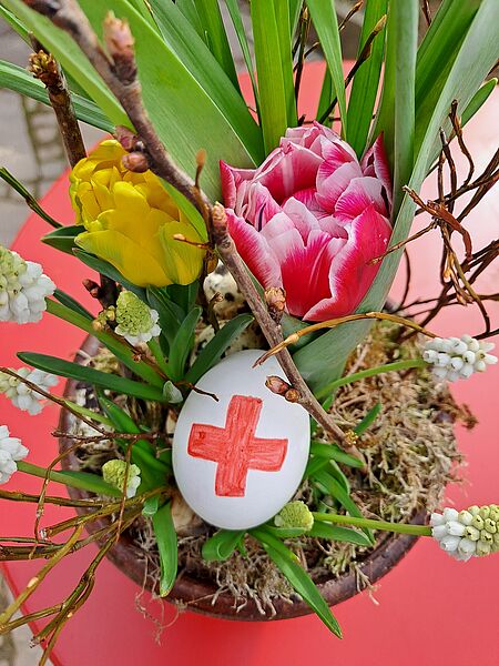 Ein mit einem roten Kreuz verziertes Osterei liegt in einem Blumengesteck.
