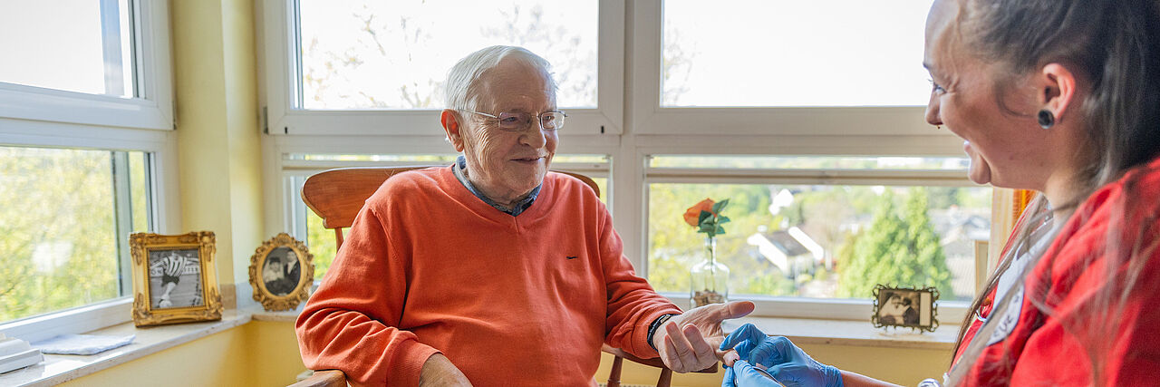Ein Senior sitzt auf einem Stuhl in seinem Zimmer im Seniorenzentrum, während eine Pflegekraft seinen Blutzucker misst.