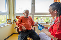Ein Senior sitzt auf einem Stuhl in einem Zimmer im Seniorenzentrum, während eine Pflegekraft seinen Blutzucker misst.
