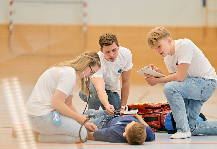 Drei Schulsanitäter versorgen ein anderes Kind in der Sporthalle. Ein Mädchen hat ein Stethoskop im Ohr, ein Notfallrucksack liegt daneben.