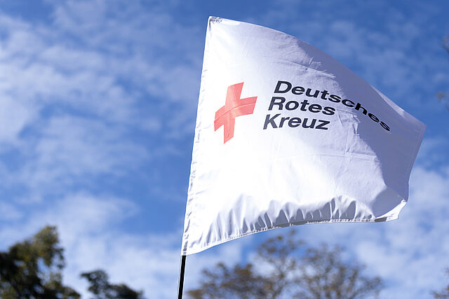 Eine weiße Fahne mit dem Logo und Schriftzug des Deutschen Roten Kreuzes weht in der Sonne.