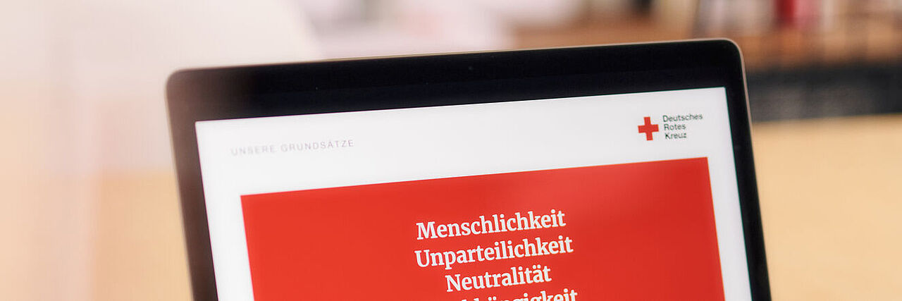 Ein Laptop-Bildschirm zeigt eine Internetseite des DRK, die auf rotem Hintergrund in weißer Schrift die Grundsätze darstellt.