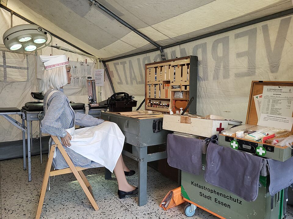 Historischer Behandlungsplatz mit einer dargestellten Einsatzkraft, medizinischer Ausrüstung und einem Wagen des Katastrophenschutzes der Stadt Essen.