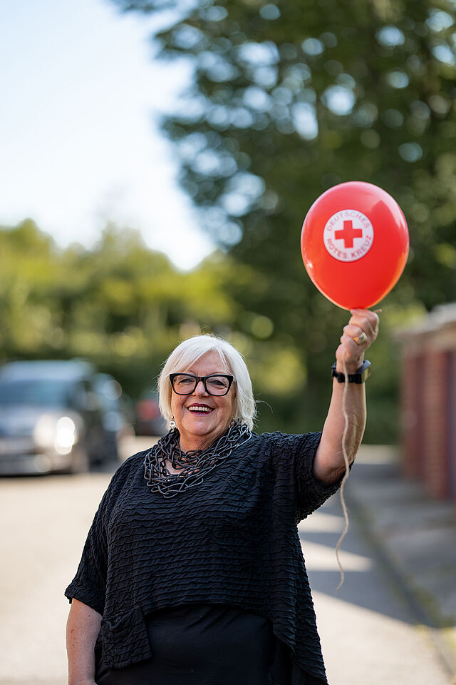 Ältere Frau hält roten Luftballon mit DRK-Logo in die Luft