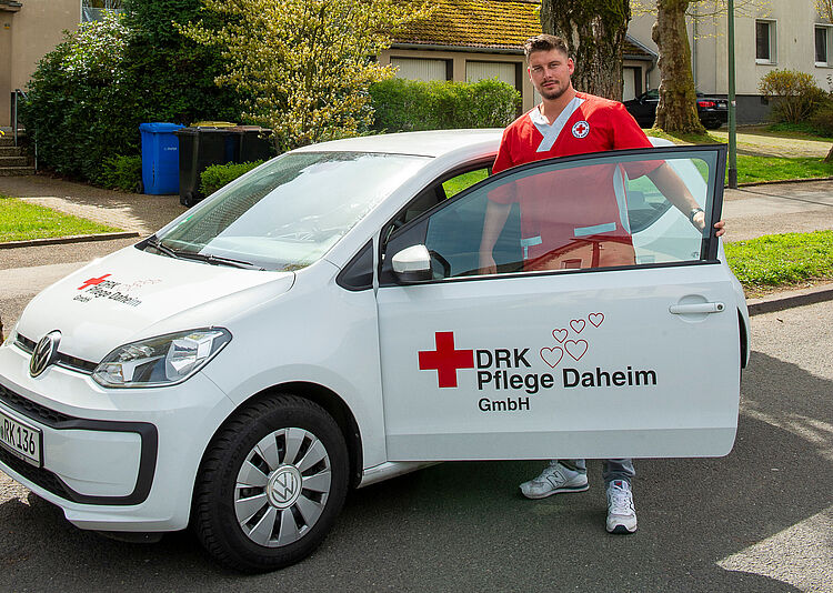 Ein Mitarbeiter in roter Dienstkleidung steht in der geöffneten Fahrertür des Autos der DRK Essen Pflege Daheim.