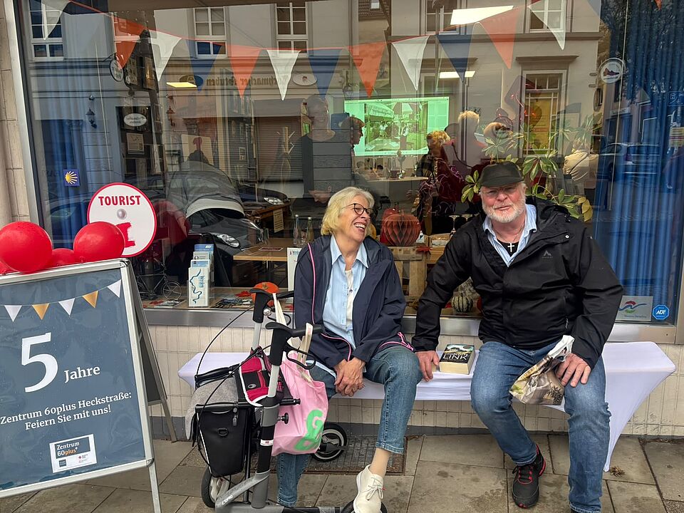 Jubiläum Zentrum 60plus Eine Seniorin und ein Senior sitzen lachend auf einer Bank vor dem Schaufenster des Zentrum 60plus in Werden.