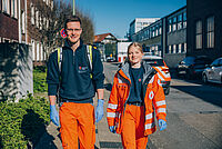 Ein junger Mann und eine junge Frau in Einsatzkleidung laufen eine Straße entlang. Im Hintergrund ist ein Einsatzfahrzeug sichtbar.