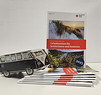 Stapel von DRK-Seniorenreisekatalogen mit einem aufrecht stehenden Exemplar, dessen Titel sichtbar ist. Ein VW-Bus-Modell steht dekorativ darauf.