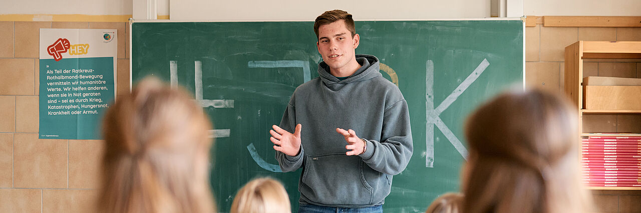 Ein älterer Schüler in Hoodie steht vor einer Tafel in einer Klasse und erklärt den Schüler*innen etwas zum JRK.