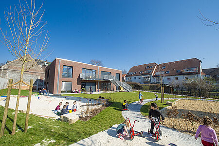 Claras Garten, Gartenansicht Das Gebäude des Kindergartens ist umgeben von einem großen Garten und Sandflächen, wo überall Kinder spielen, mit Fahrzeugen fahren und toben.