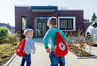Das Bild zeigt zwei Kinder von hinten, die auf das Gebäude der DRK-Kita Claras Garten zu rennen. Die tragen beide einen roten Beutel mit DRK-Logo auf dem Rücken.