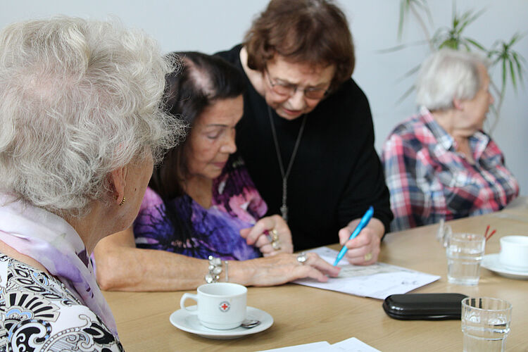 Gedächtnistraining Mehrere Seniorinnen sitzen an einem Tisch mit Kaffeetassen und Sudoku-Zetteln. Eine ältere Dame beugt sich zu einer anderen und erklärt ihr etwas.