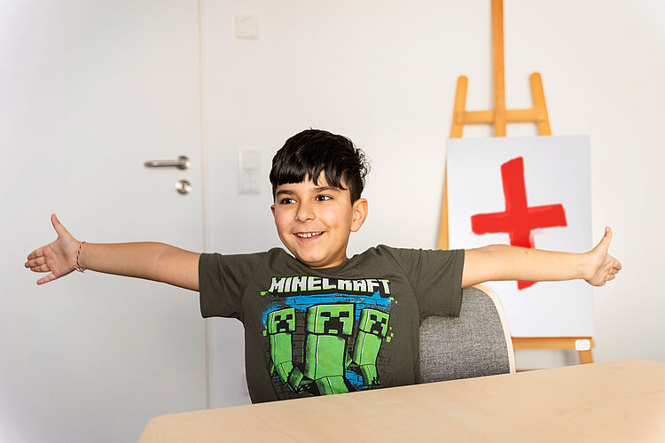 Kind Ein kleiner Junge streckt seine Arme nach rechts und links aus. Im Hintergrund steht ein Bild mit einem Roten Kreuz auf einer Staffelei.