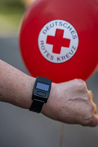 Handgelenk mit DRK-Smartwatch im Hintergrund ist ein roter Ballon mit DRK-Logo