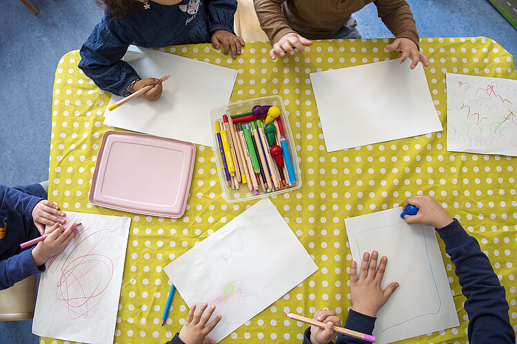 Aus der Vogelperspektive: Kinderhände malen auf weißem Papier Bilder, der Tisch ist mit einer gelben Decke abgedeckt.